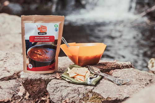 Happy Yak freeze-dried meal served outdoors on a rock, with a bowl of hot soup, crackers, and a waterfall in the background.